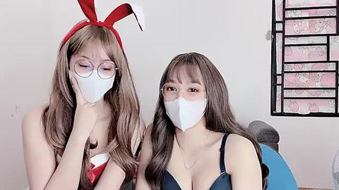 Media: Video of two Asian women in bunny costumes, wearing white face masks, sitting on a bed with a pink floral wall behind them.