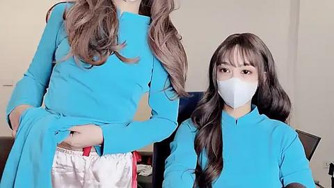 Media: Video of an Asian woman with long, wavy brown hair and pale skin, wearing a blue surgical gown and white face mask, lifting her gown to reveal white pants.