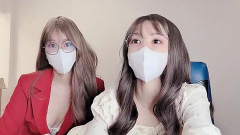 Media: Video of two East Asian women in surgical masks, one in a red blazer, the other in a white blouse, both with long, straight brown hair, sitting indoors, wearing glasses.