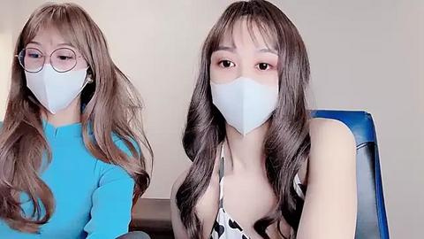 Media: A video of two young women with long, wavy brown hair and light skin, wearing blue face masks, sitting next to each other. One wears round glasses and a blue top, while the other has a black and white polka dot top.