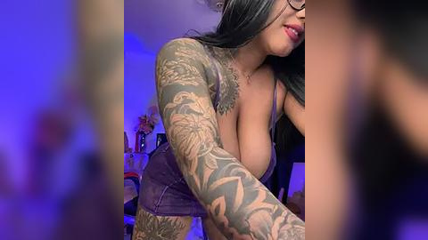 Media: A video of a voluptuous woman with long black hair, wearing a low-cut top that reveals ample cleavage, and adorned with intricate tattoos on her arms. The background features a blurred, dimly lit room with purple lighting.