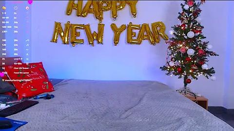 Media: Video of a festive bedroom with a bed covered in a gray quilt, \"Happy New Year\" gold balloons on the wall, a decorated Christmas tree, and a red \"New Year\" gift box.