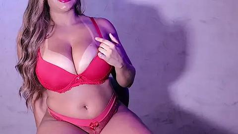 Media: Video of a voluptuous, light-skinned woman with long, wavy brown hair, wearing red lace lingerie, posing against a textured gray background. She has large breasts, a curvy figure, and red lipstick.