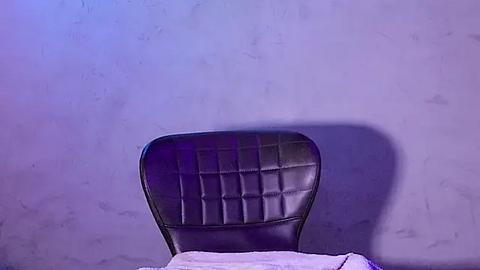 Media: A video of a black leather chair with a quilted backrest against a textured, light gray wall, bathed in soft purple lighting, creating a minimalist, modern ambiance.