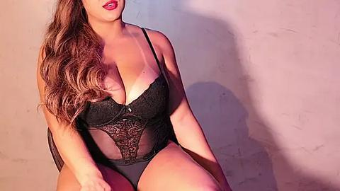 Media: Video of a curvy, light-skinned woman with long, wavy brown hair, wearing a black lace bodysuit with a plunging neckline, sitting against a textured beige wall, lips painted red.