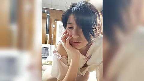 Media: A candid video captures a young Asian woman with short black hair, wearing a white top, leaning on her elbow, deep in thought. The background reveals a cozy living room with wooden furniture and a blurred figure.