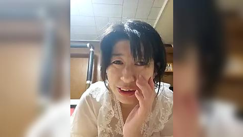 Media: Video of a young Asian woman with dark hair, wearing a white lace dress, smiling, with her finger on her cheek, in a room with a tiled ceiling and wooden paneling.