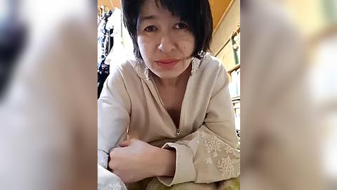 Media: Video of an East Asian woman with short black hair and fair skin, wearing a cream-colored robe, lying on a bed with a beige blanket. The background shows a blurred, warm-toned room.