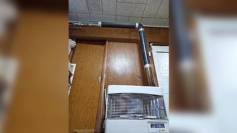 Media: A video of a small, cluttered room with a wooden paneled wall, a white air conditioner, and a black pipe running horizontally. The ceiling has a grid of fluorescent light panels.