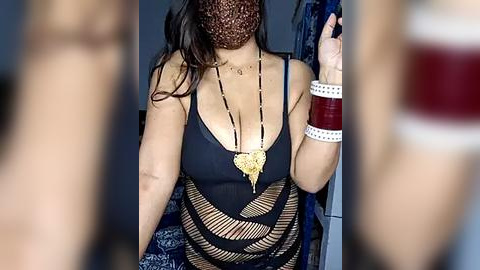 Media: Video of a woman with medium skin tone wearing a black, low-cut, fishnet top, a gold heart pendant necklace, and a red mask, with a blurred background of blue and red patterns.