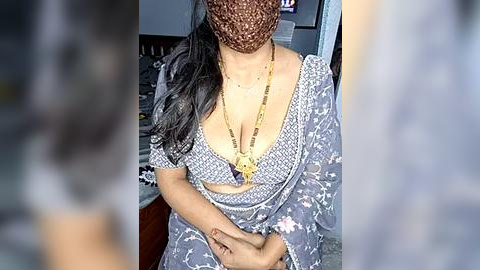 Media: A video of a woman in a blue floral sari with a low-cut blouse, gold necklace, and brown face mask, standing indoors with blurred background.