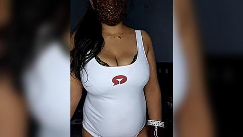 Media: Video of a woman wearing a tight, white tank top with a red logo and a face mask, standing indoors with blurred background.