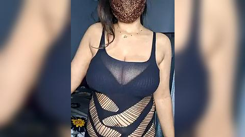 Media: Video of a woman in a sheer black mesh top revealing her large breasts, with her face obscured by a brown lace mask. Background shows a blurred room with a flowered pattern.