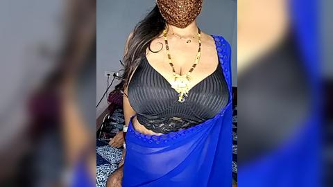 Media: Video of a woman in a blue saree with a black lace bra, gold necklace, and brown headscarf, blurred background.