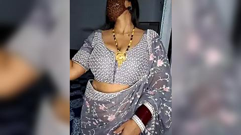 Media: Video of a woman wearing a gray saree with floral patterns, a golden pendant, and a maroon bindi. The background is blurred, focusing on the woman's attire and accessories.