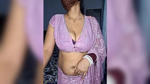 Media: Video of a South Asian woman in a pink sari with a plunging neckline, showcasing medium-sized breasts, wearing a headscarf, and a beaded necklace. Background shows a dark room with a patterned rug.