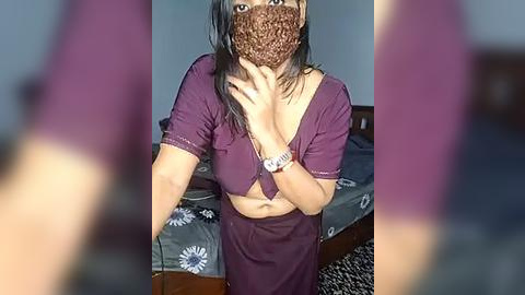 Media: Video of a woman with medium-dark skin, long dark hair, and a brown face mask, wearing a purple crop top and skirt, standing in a bedroom with a bed and patterned blanket.