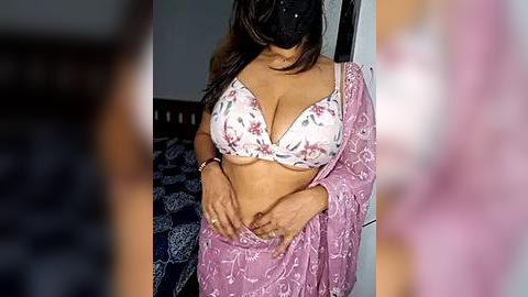 Media: Video of a woman with medium brown skin and long, straight brown hair, wearing a white bra with floral pattern and a pink, floral-patterned saree. She is adjusting her saree in a dimly-lit bedroom with a blue-patterned bedspread in the background.