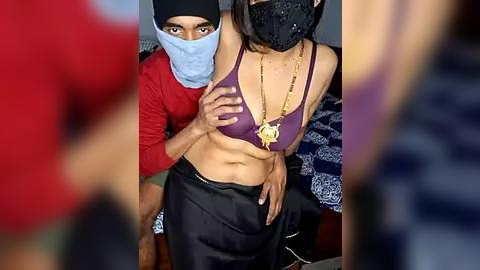 Media: Video of a South Asian woman with medium skin tone, wearing a purple bra, black mask, and black pants, being held by a man in a red shirt and black beanie. Background shows a bed with blue and white patterned sheets.