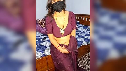 Media: A video of a woman in a purple saree, black mask, and gold necklace, sitting on a bed with blue and white patterned sheets, in a dimly lit bedroom.