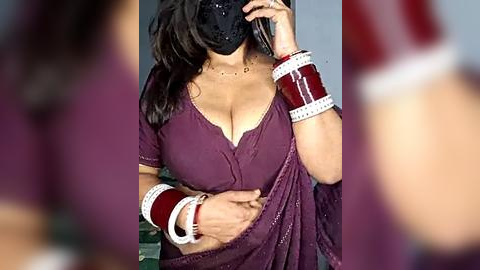 Media: Video of an Indian woman in a maroon sari, holding her belly, wearing a black bindi, red bangles, and a necklace. Background is blurred, focusing on her attire and accessories.