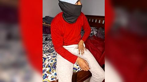 Media: Video of a person in a red long-sleeve shirt, white pants, and black mask sitting on a bed with a colorful patterned duvet. Background includes a wooden headboard and red curtains.