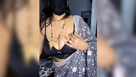 Media: Video of a woman with medium skin tone wearing a black bra, gold necklace, and a black mask, seated in a blurred room, partially visible.