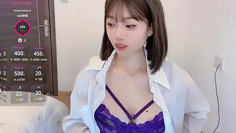 Media: Video of an Asian woman with fair skin and straight brown hair, wearing a white button-down shirt, revealing a purple lace bra. Background shows a digital clock and a wall.