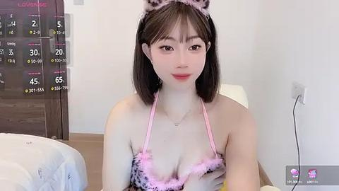 Media: Video of an East Asian woman with fair skin, shoulder-length dark hair, and wearing a pink and white cat-themed lingerie set. She sits on a bed in a minimalist room with a wooden floor and white walls.