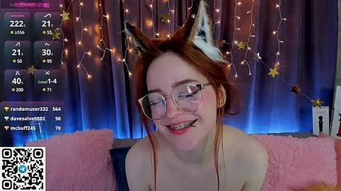 Media: Video of a young woman with red hair, wearing cat ears and glasses, grinning with braces, on a pink couch, in a cozy room with fairy lights.