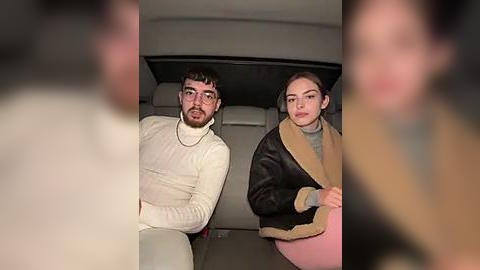 Media: A video of a man with glasses and beard wearing a white turtleneck, sitting next to a woman with brown hair in a beige coat. They are in a car at night.