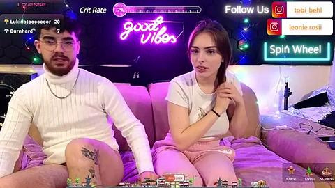 Media: Video of a man with a beard and glasses, and a woman with long hair, both wearing white sweaters and pink pants, sitting on a bed, surrounded by a neon-lit room with a \"Good Vibes\" sign.