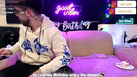 Media: Video of a man with a beard, glasses, and a white hoodie, sitting on a couch, watching a birthday stream. Background includes a neon sign, a cake, and social media icons.