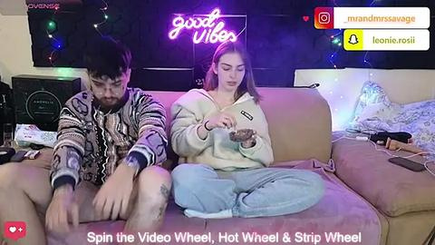 Media: Video of two people on a beige couch in a dimly lit room with a neon \"good vibes\" sign and a cluttered background.