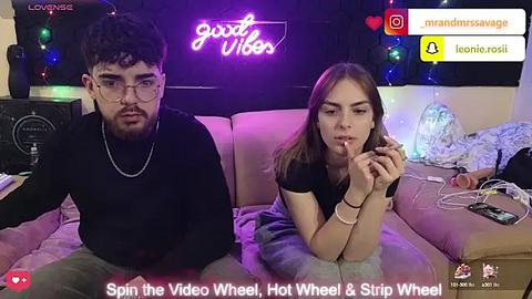 Media: Video of a young couple in a dimly lit room with neon lights. The man has a beard and wears a black sweater, while the woman has long hair and is smoking a cigarette.