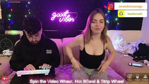 Media: Video of a young man with glasses, beard, and black hoodie playing a video game, and a young woman with shoulder-length brown hair in a black tank top and gray shorts, both sitting on a beige couch in a dimly lit room with colorful string lights and a \"Good Vibes\" neon sign.