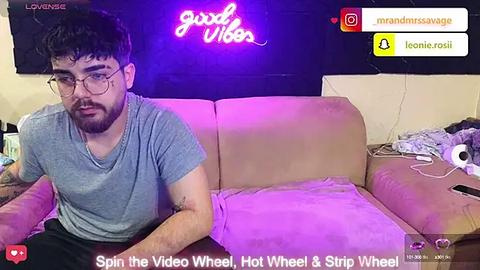 Media: Video of a young man with glasses and a beard, sitting on a beige couch with purple lights and a neon sign behind him, wearing a gray t-shirt.