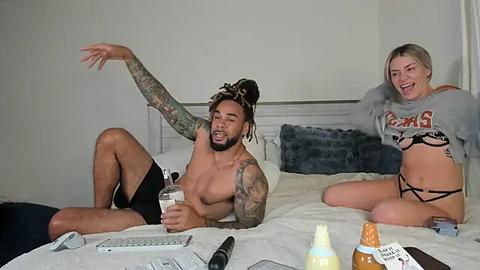 Media: Video of a shirtless, tattooed man with dreadlocks and a woman with blonde hair, both laughing on a bed in casual attire, surrounded by personal items.