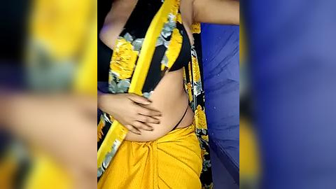 Media: Video of a South Asian woman wearing a black floral top and yellow dhoti, holding her pregnant belly. Background features blurred figures in yellow and blue clothing.