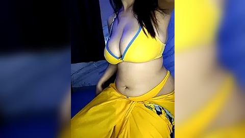 Media: Video of a woman in a yellow bikini top and matching sarong, sitting on a bed. Her long dark hair falls over her shoulders, and the background is dimly lit with blue and yellow tones.