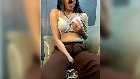 Media: Video of a young woman with medium skin tone and long dark hair, wearing a beige bra, brown pants, and a distressed patterned bag, sitting on a beige couch, mouth open in surprise.