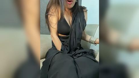 Media: A video of a South Asian woman with long, dark hair, wearing a black, sleeveless top and draped sari, sitting on a beige couch, hands in her lap, and a bracelet on her wrist.