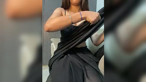 Media: Video of a woman with medium skin tone and long dark hair, wearing a black dress, adjusting the top, revealing a black bra, seated on a white chair in a modern room.