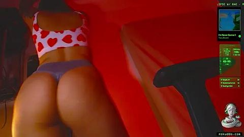 Media: Video of a woman with dark skin wearing a white heart-patterned crop top and gray thong, captured from behind on a red background, with a gaming interface in the corner.