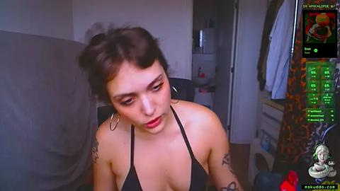 Media: A video of a young woman with short dark hair, tattoos, wearing a black halter top, sitting indoors with a gaming screen displaying a character in the background.