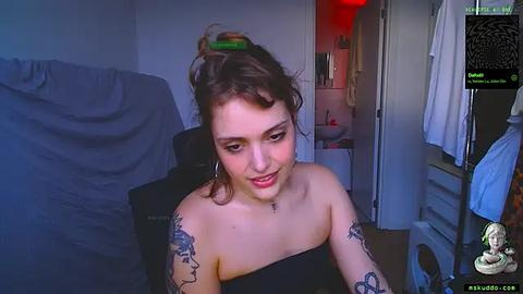 Media: Video of a young woman with light skin and short brown hair, smiling, wearing a black strapless top, with tattoos on her arms, sitting in a dimly lit room with a TV screen displaying an anime character.