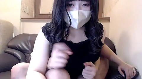 Media: Video of an Asian woman with long black hair, wearing a white mask, black lace top, and a white dress, sitting on a brown leather couch in a beige room.