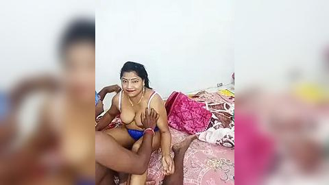 Media: A blurred video of a South Asian woman with medium skin tone and dark hair, wearing a blue bra, sitting on a pink-patterned bed with a man's arm around her, in a cluttered room.