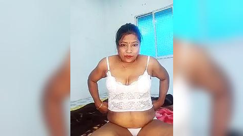 Media: A video of a plus-sized woman with medium brown skin and dark hair tied back, wearing a white lace bra and panties, sitting on a bed with a window behind her.