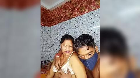 Media: Video of an Indian woman with medium skin tone, dark hair in a ponytail, wearing a white lace bra, sitting on a bed with a young man wearing a blue mask, in a room with red floral wallpaper and white mosaic tiles.